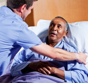 Male nurse (30s) helping African American senior man (60s) in hospital bed.