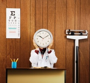 Caucasian male doctor sitting at desk holding clock over face.